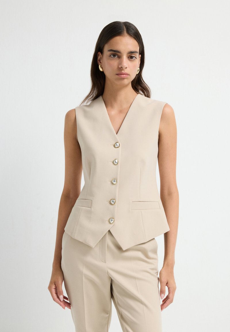 Blazer sans manches beige avec un décolleté en V plongeant, fermeture à cinq boutons à l'avant et deux poches latérales ; tissu lisse avec une coupe ajustée.
