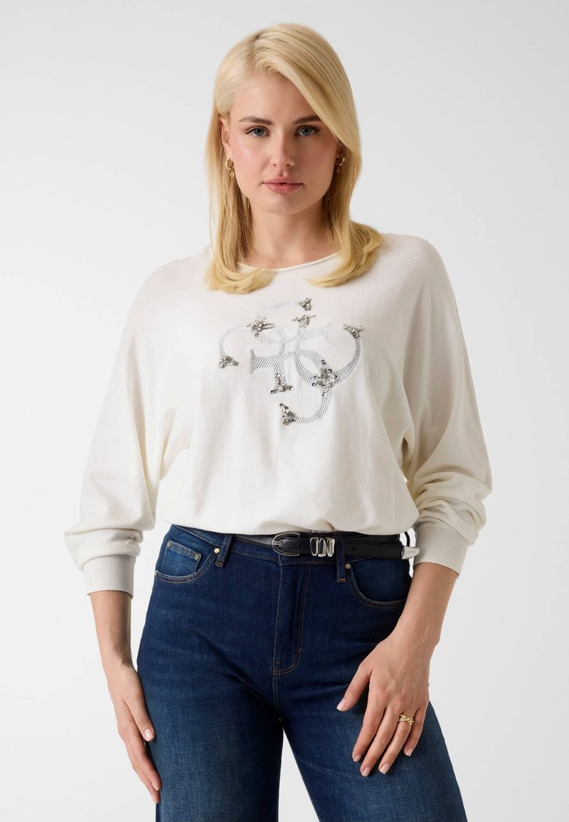 Femme aux cheveux blonds portant un haut blanc à manches longues avec des ornements décoratifs et un jean bleu foncé avec une ceinture noire.