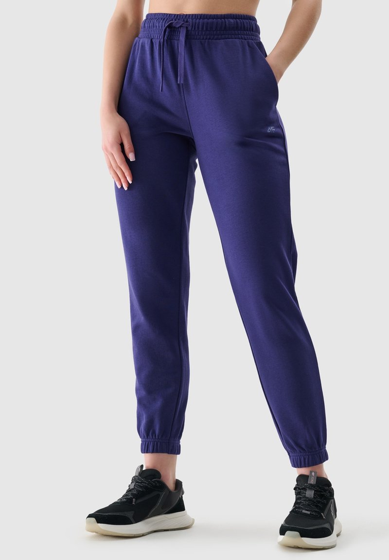 Fioletowe dresy z elastycznym paskiem w talii i ściągaczami, z bocznymi kieszeniami i miękką fakturą, w połączeniu z czarnymi butami sportowymi.