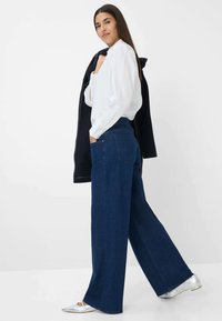 Brede, donkerblauwe spijkerbroek met hoge taille, gecombineerd met een witte blouse met lange mouwen. Er worden zilveren platte schoenen gedragen.