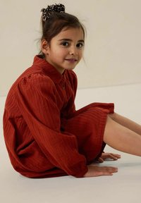 Rotes geripptes Kleid mit langen Ärmeln und Kragen, mit Knopfverschluss. Das Kind sitzt auf dem Boden, mit einem gemusterten Haargummi im Haar.