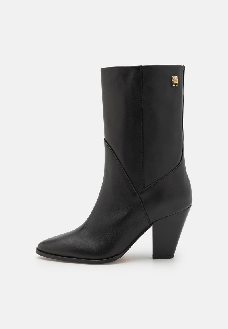 Musta nahkainen nilkkuri, jossa on terävä kärki, teksturoiduilla yksityiskohdilla, kulmikas muotoilu ja keskikorkea kartiomainen korko. Mukana kultainen logo.