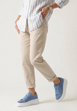 Person trägt beige hochgekrempelte Hose, blaue Turnschuhe mit weißen dicken Sohlen und ein langärmeliges gestreiftes Hemd, steht mit einem angehobenen Bein.