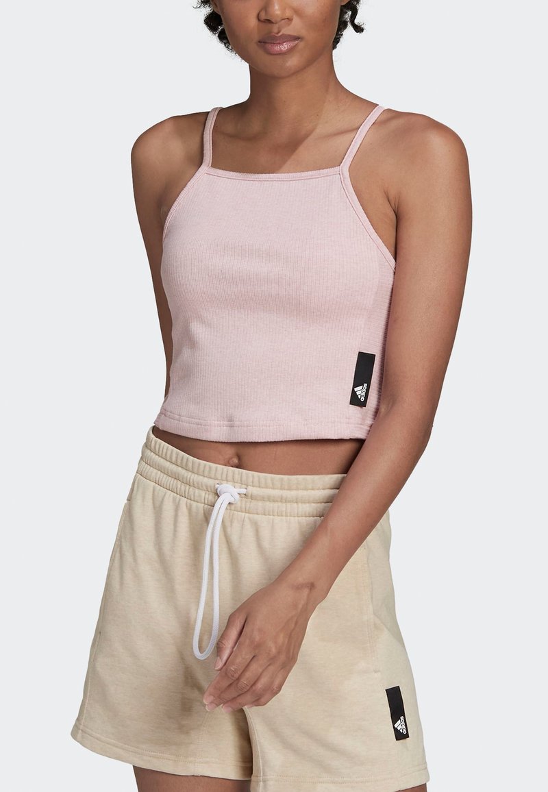 Top corto de canalé en rosa claro con tirantes finos, que presenta una etiqueta de logo negra en el lateral. Combinado con shorts beige y cordón blanco.