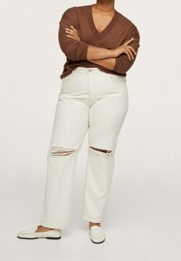 Jeans blancs déchirés avec une coupe décontractée et une taille haute, associés à un pull en V marron et des mocassins de couleur claire.