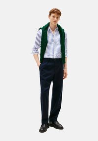 Camicia a righe con maniche lunghe, gilet in maglia verde appoggiato sulle spalle, pantaloni eleganti scuri e scarpe nere.
