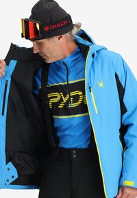 Giacca da sci impermeabile blu con interno nero, dettagli gialli e logo di un ragno. Design con colletto alto, tasche con zip e polsini regolabili.