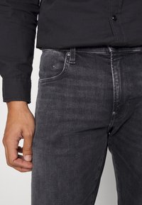 Vaqueros de mezclilla negros con un acabado desgastado, diseño de cinco bolsillos y herrajes plateados. La textura parece suave con detalles de costura visibles.