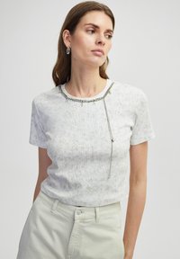 T-shirt blanc à manches courtes avec un motif texturé, présentant un détail de chaîne en argent autour du cou et une chaîne d'accent qui pend.