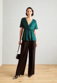 Top avvolgente teal con maniche corte, caratterizzato da una leggera lucentezza e un motivo testurizzato. Abbinato a pantaloni larghi neri e tacchi a punta.