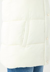 Leichter, cremefarbener Puffer-Westen mit horizontalen Nähten, einer Seitentasche und goldfarbenen Knöpfen zum Schließen. Weiche, gesteppten Textur.
