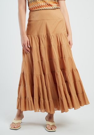 Woman wearing a long, tiered tan skirt, multicolored knit top, and cream open-toe sandals standing against a plain background.