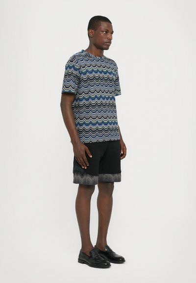 Haut à manches courtes à motifs avec un design ondulé bleu et vert ; associé à un short noir avec un ourlet en zigzag. Mannequin portant des chaussures noires.