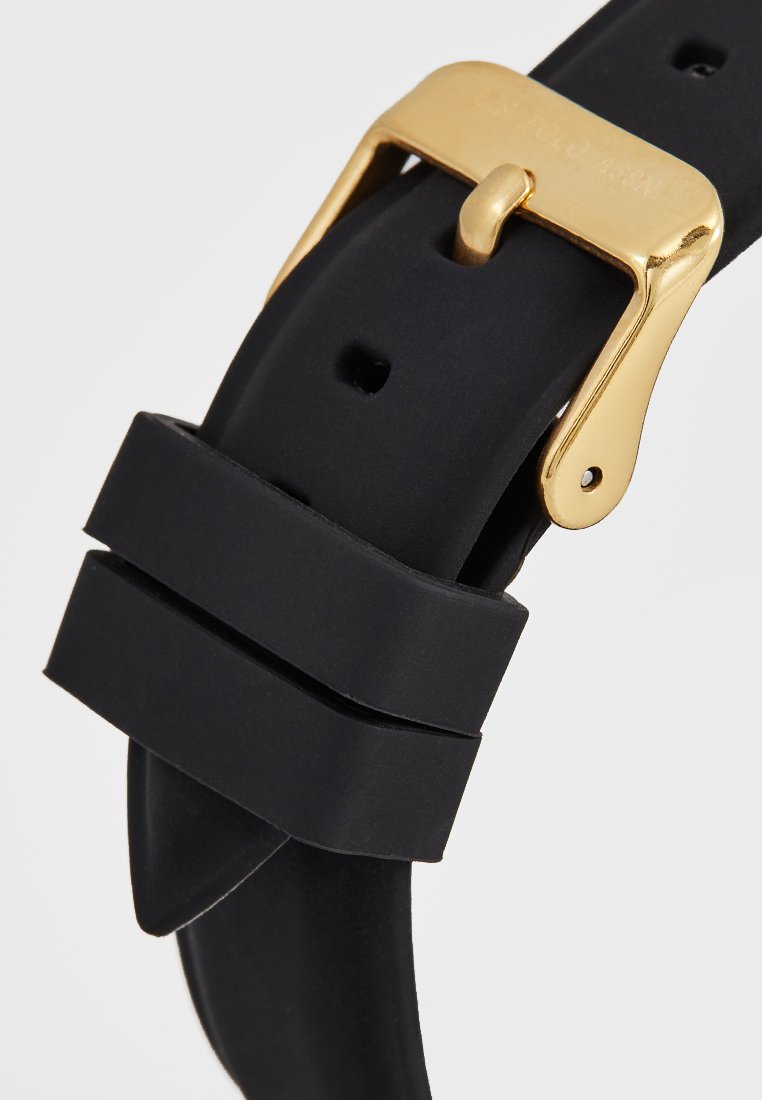 Correa de reloj de goma negra con hebilla de metal dorado. Presenta dos acentos estriados y múltiples agujeros para ajustabilidad. Textura suave en todo.