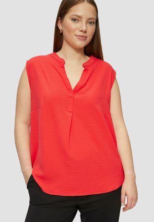 Blouse sans manches rouge avec un col en V et une coupe décontractée, dotée d'une finition texturée subtile et d'un ourlet arrondi. Associée à un pantalon noir.