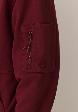 Burgundy sweatshirt with a zippered arm pocket, textured fabric, and embroidered logo patch on the pocket. Close-up view of the sleeve.