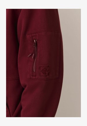 Bordeaux sweatshirt met een ritszak op de mouw, textuurstof en geborduurd logo op de zak. Close-up van de mouw.