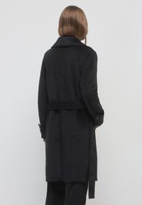 Donna con lunghi capelli castani che indossa un cappotto nero con cintura, in piedi di spalle su uno sfondo chiaro e semplice.