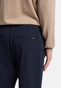 Marineblaue Hose mit glatter Textur, verfügt über eine Gesäßtasche und dezentes Branding. Kombiniert mit einem hellbeigen Pullover.
