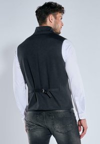 Dunkelgraue Weste mit hohem Kragen, maßgeschneidertem Design und Rückenriemendetail, getragen über einem weißen Langarmshirt und dunklen Jeans.