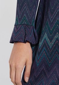Abito a motivo a zigzag con una combinazione di colori viola e turchese. Presenta dettagli con volant sui polsini e una finitura in tessuto testurizzato.
