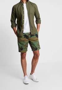 Chemise boutonnée vert olive associée à un short camouflage vert et noir, complétée par des baskets blanches. La chemise possède une poche poitrine.