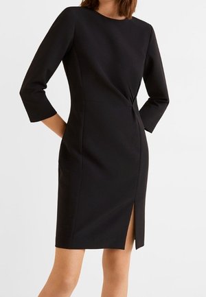 Femme portant une robe noire cintrée au genou avec des manches trois-quarts et une fente à l'avant, debout devant un fond uni.