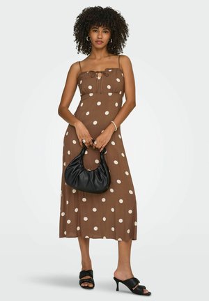 Mujer con un vestido midi marrón con lunares blancos, llevando un bolso negro y sandalias negras de tacón.