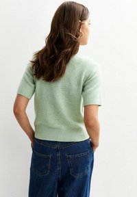 Maglione verde chiaro a maniche corte con vita aderente, realizzato in materiale morbido. Indossato con jeans blu in denim a vita alta con tasche posteriori.