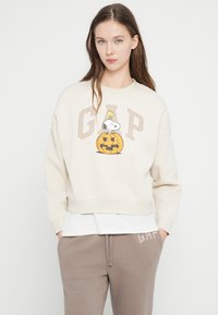 Młoda kobieta ubrana w beżową bluzę GAP z nadrukiem Snoopy'ego i Woodstocka na rzeźbionej dyni, w połączeniu z taupe'owymi spodniami dresowymi GAP.