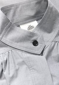 Lys grå skjortekrave med en sort knap fastgjort på en flap, synlige sømme og en lille hvid tøjlabel indeni kraven.