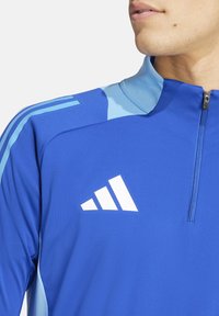 Blauwe sportieve jas met een kwart ritssluiting, lichtblauwe accenten en drie witte diagonale strepen als logo op de linkerborst. Glad textuur.