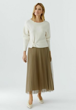 Frau trägt einen weißen langärmeligen Pullover mit Knotendetail, einen olivgrünen plissierten Midi-Rock und weiße spitze High Heels, steht vor weißem Hintergrund.