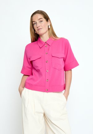 Frau trägt ein leuchtend pinkes Kurzarm-Hemd mit Taschen und cremefarbene, hoch taillierte Hose, steht mit den Händen in den Taschen.