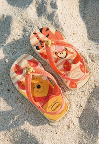 Petites tongs orange pour enfant avec des motifs Winnie l'Ourson et Tigrou reposant sur un sol sablonneux clair.