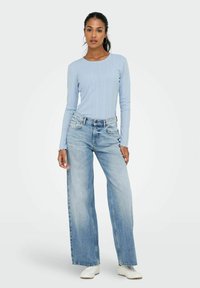 Das Model steht selbstbewusst in einem hellblauen, gerippten Oberteil und weiter geschnittenen Jeans, kombiniert mit weißen Sneakern, vor einem minimalistischen Hintergrund.
