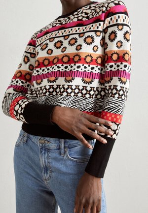 Persoon draagt een meerkleurige trui met geometrische en bloemmotieven, gecombineerd met een blauwe spijkerbroek, tegen een effen lichte achtergrond.