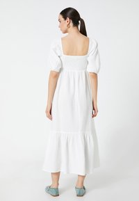Vestido blanco de mangas abullonadas con un cuerpo fruncido y dobladillo en capas. Tejido suave y texturizado. Se lleva con zapatos azules. Vista trasera mostrada.