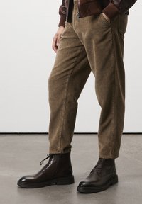Des jambes portant un pantalon en velours côtelé marron et des bottines en cuir brun foncé, avec une main dans la poche, se tenant sur un sol gris.
