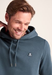 Blue hooded sweatshirt made from cotton blend fabric. Features a drawstring, embroidered logo on chest, and a soft texture.