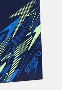 Ναυτικό μπλε ύφασμα με αφηρημένα γεωμετρικά σχέδια σε πράσινο λάιμ και μπλε, και το μπλε λογότυπο Speedo τυπωμένο κοντά στο τελείωμα.