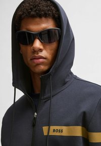 Mörkgrå zip-up hoodie med en gyllene horisontell rand och "BOSS"-logotyp. Modellen bär svarta wrap-around solglasögon. Slät textur.