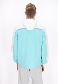 Giacca leggera in azzurro chiaro e bianco, con cappuccio, maniche lunghe e striscia riflettente sulla schiena. Tessuto morbido.
