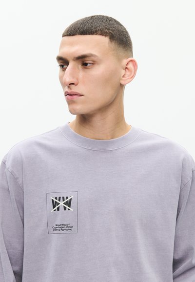 Jeune homme aux cheveux courts et foncés portant un sweat-shirt violet clair avec un logo "WW" barré et du texte sur le côté gauche de la poitrine.