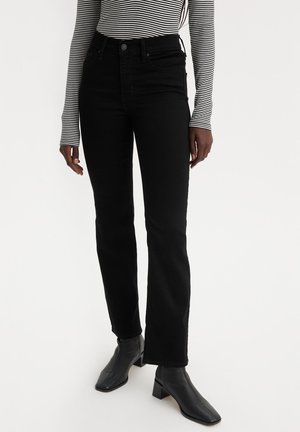 Vrouw draagt zwarte jeans met rechte pijpen, zwarte enkellaarsjes met hak en een wit-zwarte gestreepte top met lange mouwen.