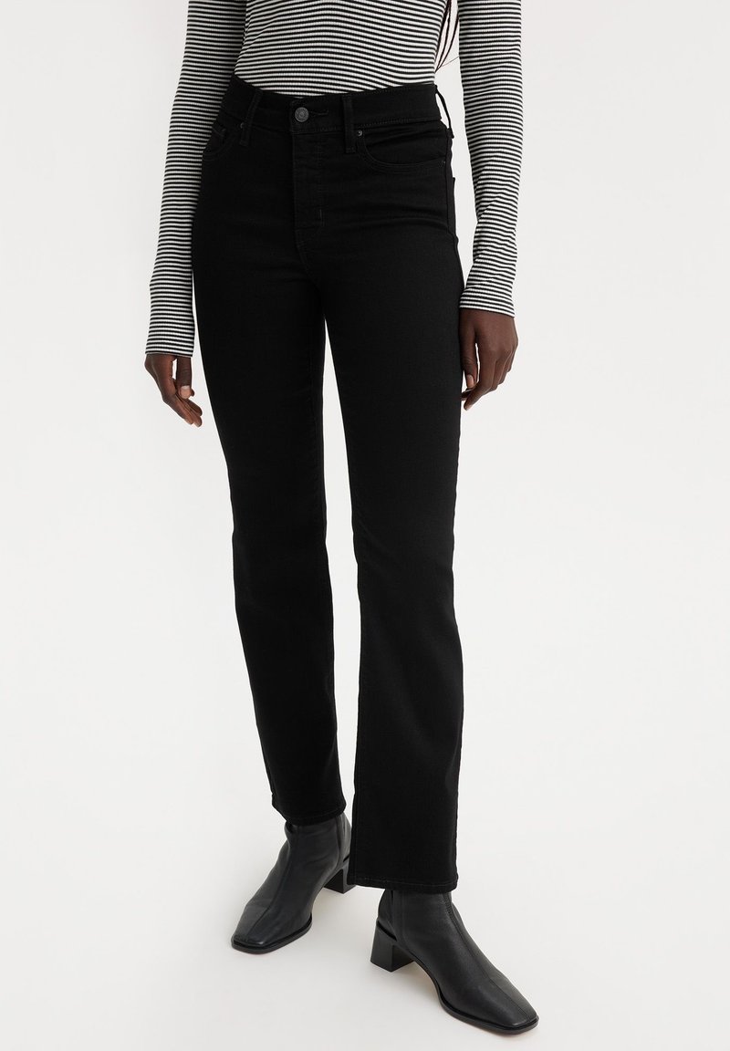 Femme portant un jean noir droit, des bottines noires à talons et un haut à manches longues rayé blanc et noir.