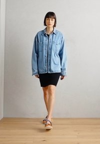 Giacca in denim oversize di colore blu chiaro con due tasche frontali, abbinata a un vestito aderente nero e sandali platform beige. Texture liscia.