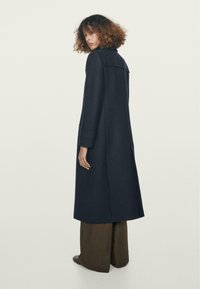 Cappotto lungo blu navy con colletto e polsini arrotolati, realizzato in tessuto testurizzato. Il design presenta una linea dritta, tasche laterali e una apertura sul retro.