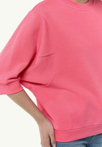 Różowy sweatshirt wykonany z miękkiego materiału. Posiada okrągły dekolt, opadające ramiona oraz luźne rękawy. Gładka tekstura bez widocznych wzorów.