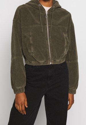 Personne portant un sweat à capuche court en velours côtelé vert olive avec fermeture éclair et cordons, ainsi qu’un pantalon noir taille haute, debout devant un fond uni.